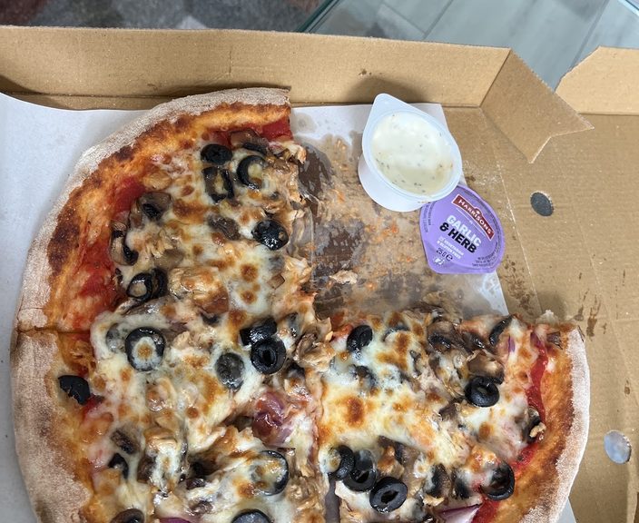 An olive, pepperoni and red onion pizza in a box with a dip pot